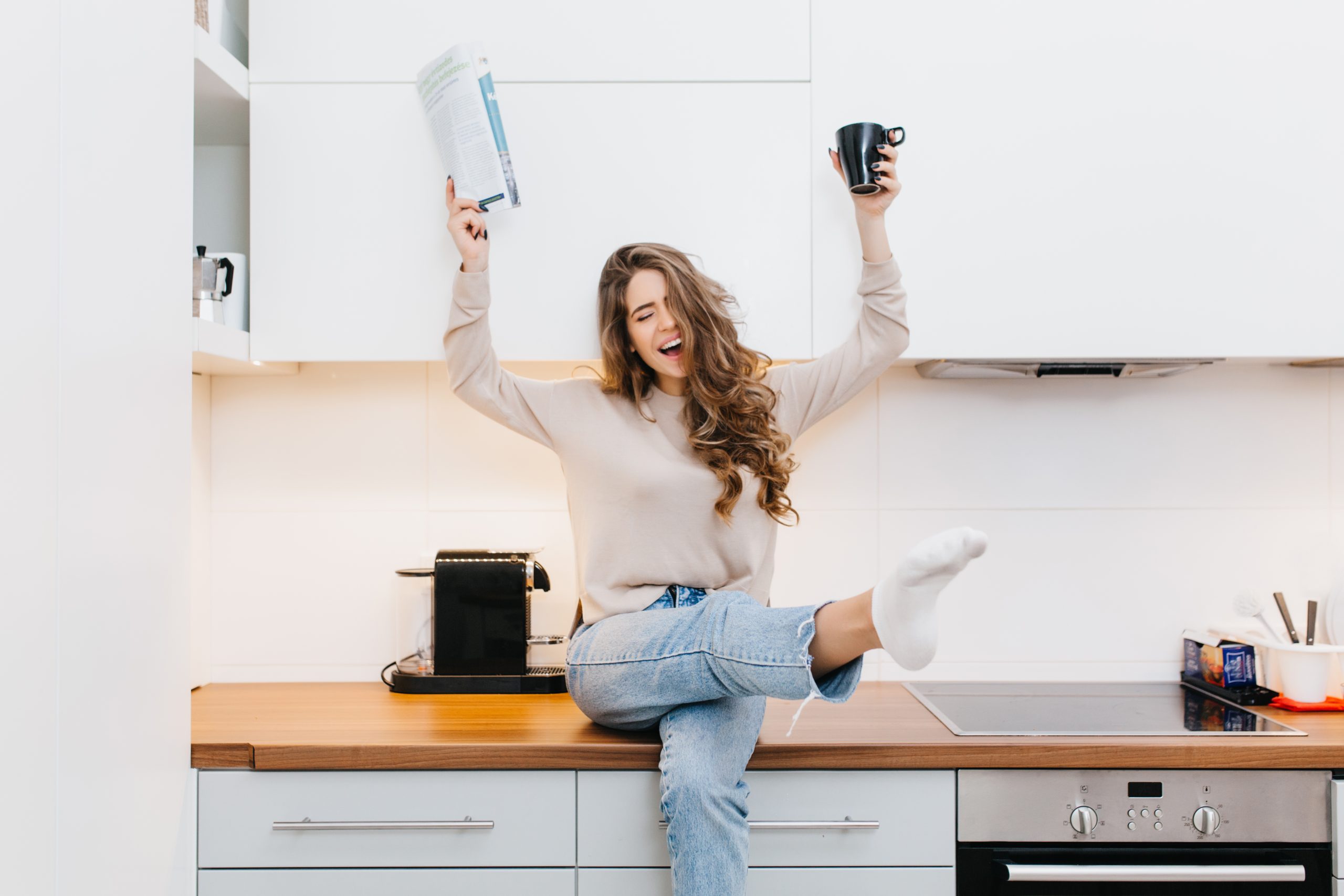 Graceful Caucasian girl wears jeans enjoying good morning in her kitchen. Indoor portrait of emotional female model drinking coffee and reading magazine..
