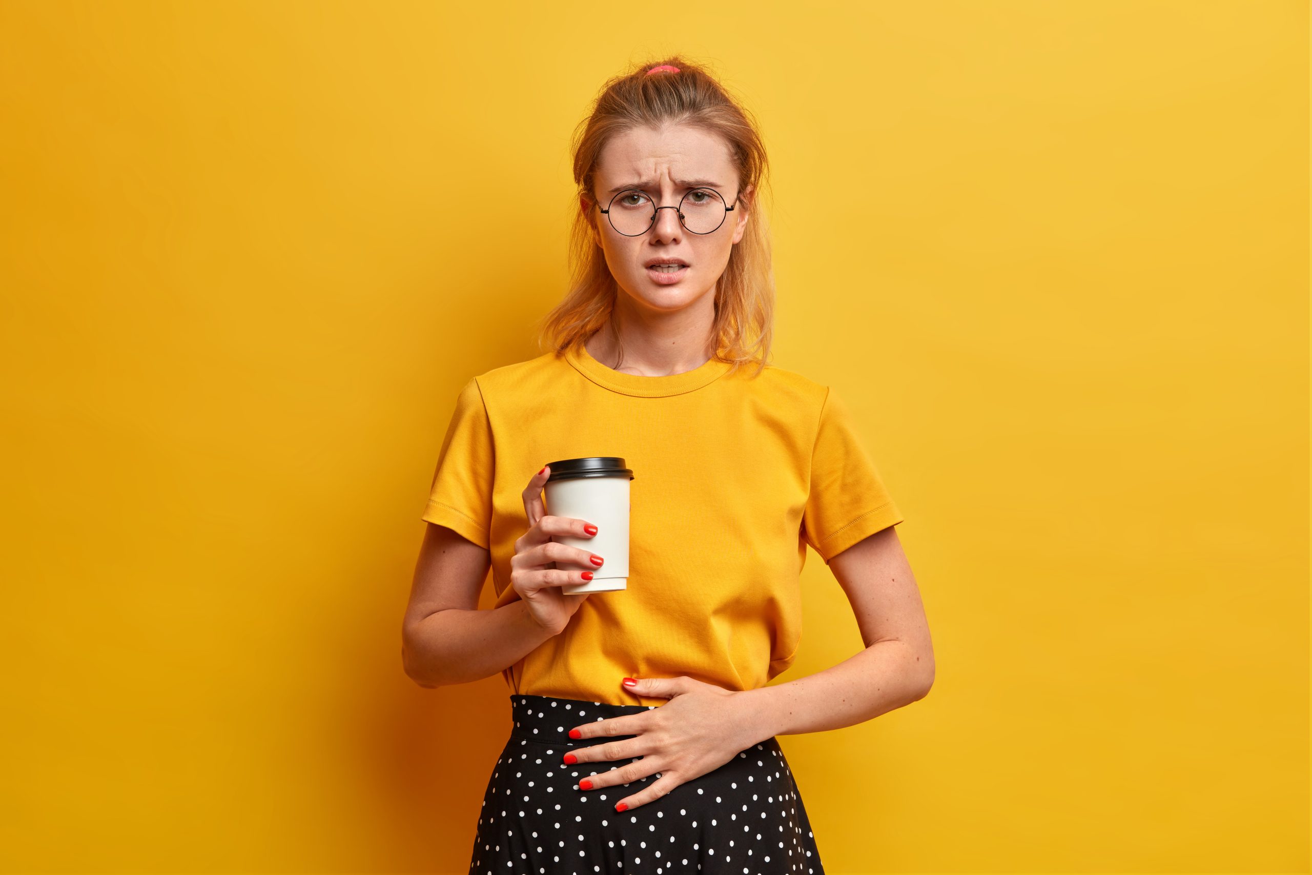 Unhappy displeased female frowns face, feels unwell, keeps hand on belly, drinks takeaway coffee, ate spoiled food, wears transparent glasses, casual yellow t shirt, stands indoor. Abdominal pain