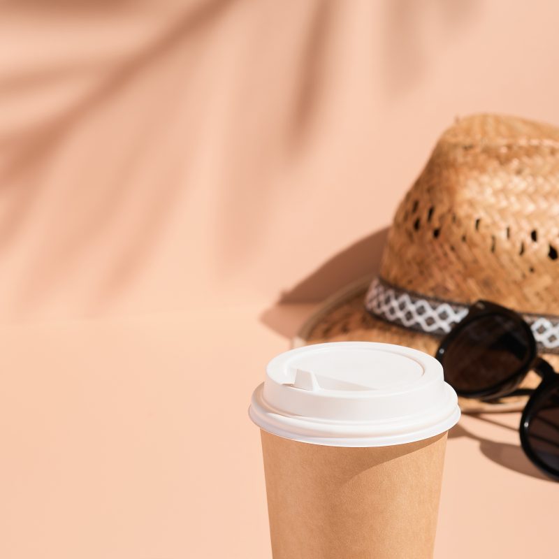 Coffee cup, straw hat and sunglasses on a light beige background shaded by palm leaves, vertical frame. Dreaming about summer and fun, enjoying life, vacations, holidays, travel, tourism concept
