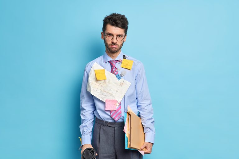 Sad unshaven man looks seriously at camera holds coffee and papers being tired of preparing report dressed in formal clothes comes to business meeting isolated over blue background works in office