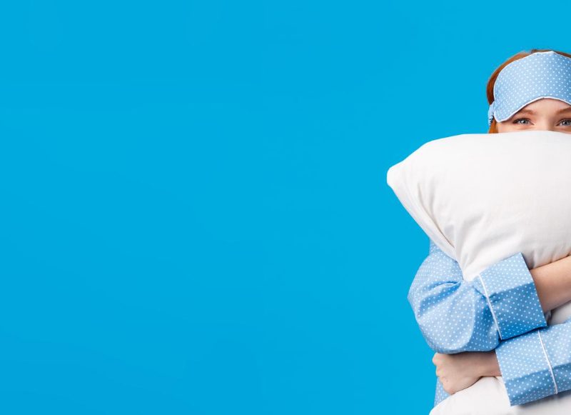 Woman in front of a blue wall holding a pillow and wearing a sleep mask