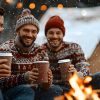 Friends in matching Christmas sweaters sitting by a campfire with a hot drink