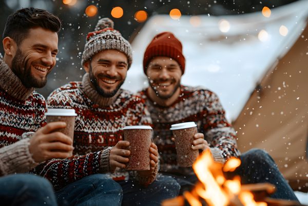 Friends in matching Christmas sweaters sitting by a campfire with a hot drink