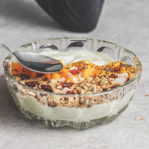 Close-up of hand holding spoon eating mango yogurt with granola