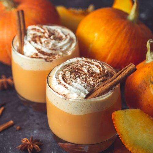 2 clear glasses with lattes and a cinnamon stick with some small pumpkins