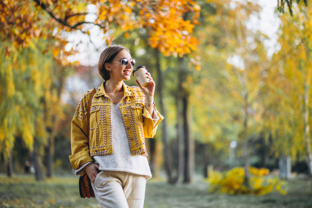 A woman wearing sunglasses and a yellow jacket walking outside with fall leaves while enjoying a drink